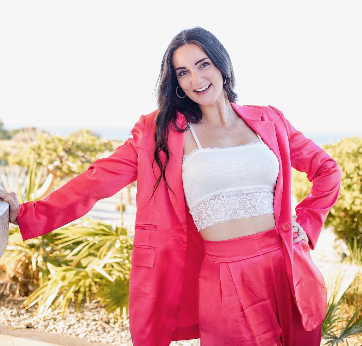 Ashley Brasseaux smiling in a hot-pink blazer and white lace bralette, hands on her hips, with palm plants and bright sky behind her