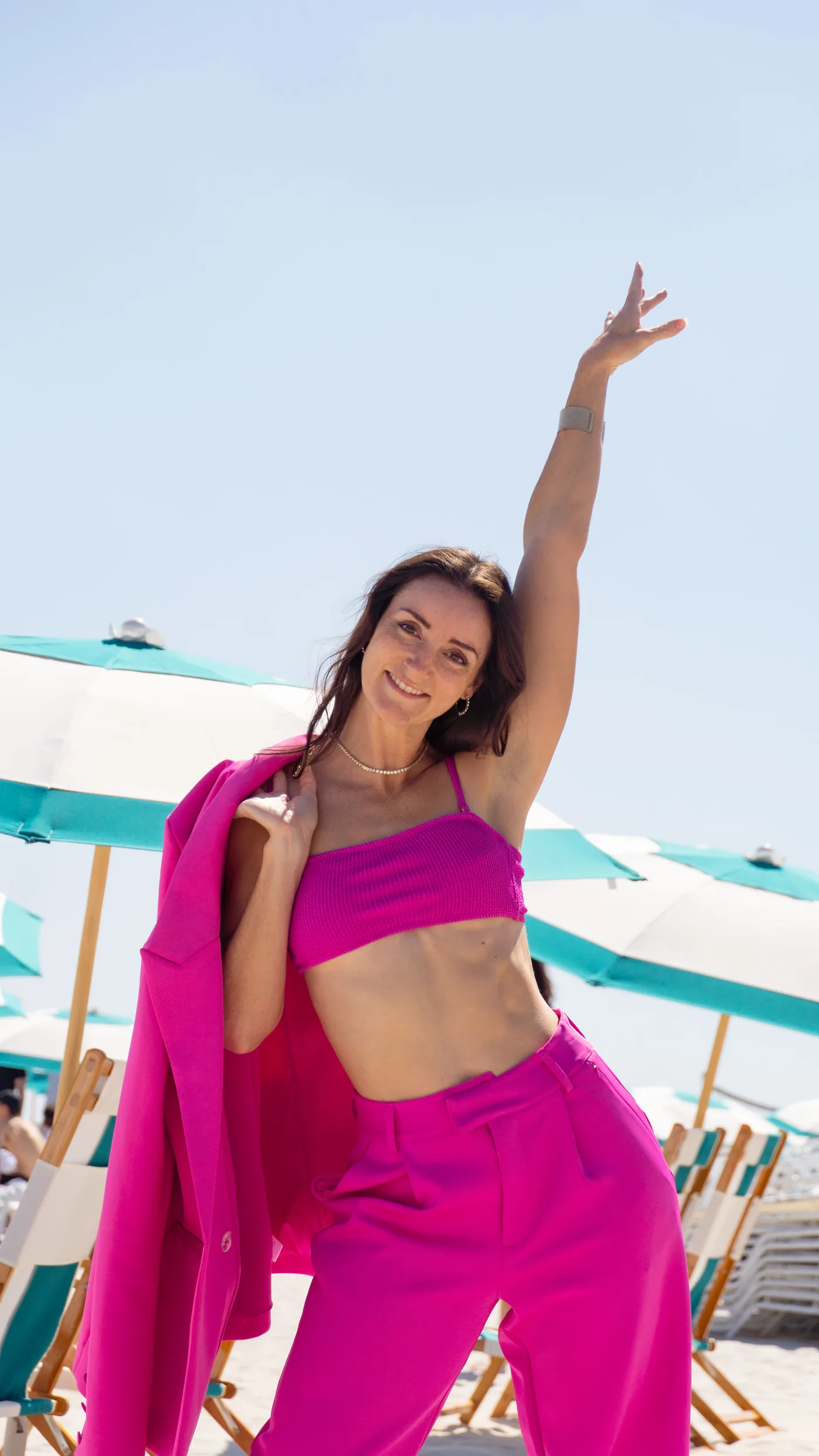 Ashley Brasseaux on a beach in a hot-pink suit and crop top, one arm raised, smiling, with teal-and-white striped umbrellas and beach chairs behind her