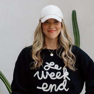 Gemma Bonham-Carter, smiling in a white baseball cap and a black sweater that reads 'Le Week End,' standing next to a cactus