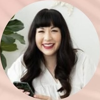 Nicole Benson, laughing in a white top with bangs and long dark hair, holding a phone, with a green plant behind her