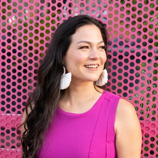 Selena Soo, smiling in a hot-pink sleeveless top with large white tassel earrings, against a coral perforated wall